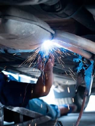 Mechanic welding exhaust system under vehicle, sparks flying, emphasizing exhaust and muffler repair services at Route 40 Auto.